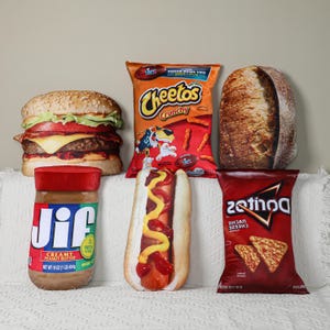 May include: A collection of food-shaped pillows, including a hamburger, a bag of Cheetos, a loaf of bread, a jar of Jif peanut butter, a hot dog, and a bag of Doritos. The Jif jar is labeled "NET WT 16 OZ (1 LB) 454g".
