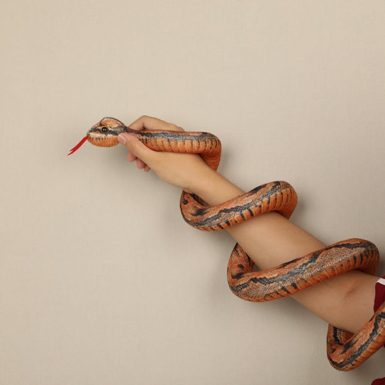 May include: An orange and black snake toy with a red tongue is wrapped around an arm. The snake's body is orange with black stripes. The snake's head is held by a hand.