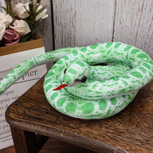 May include: A plush green and white snake toy coiled on a wooden surface. The snake has black eyes and a red tongue. The toy is designed with a scale pattern.