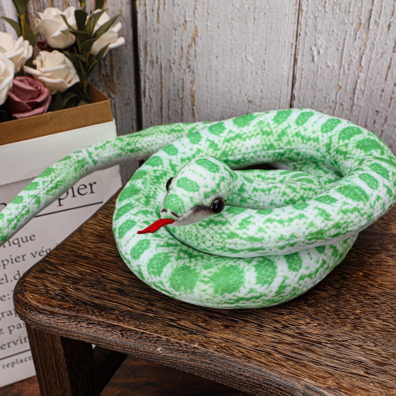 May include: A plush green and white snake toy coiled on a wooden surface. The snake has black eyes and a red tongue. The toy is designed with a scale pattern.