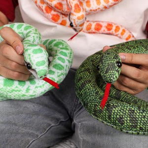 May include: Close-up of several plush snake toys in various colors. One is green with white spots, another is green with black spots, and the third is orange with white spots. Each snake has a red tongue and black eyes.