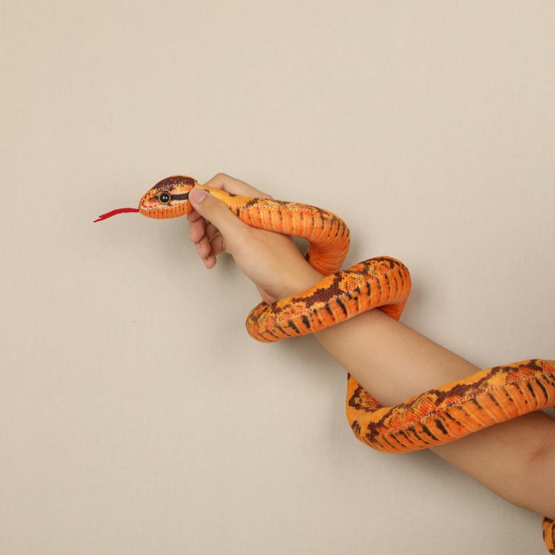 May include: An orange and brown plush snake toy with a red tongue. The snake is wrapped around an arm and hand. The snake's head is held by a hand, and the body is wrapped around the arm. The snake has black stripes.