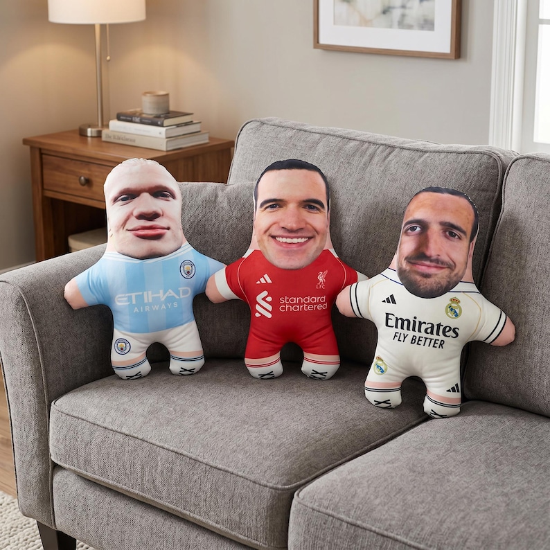 May include: Three plush dolls shaped like figures, each with a different face printed on the front. The dolls are wearing soccer jerseys in light blue, red, and white. The dolls are on a gray couch.