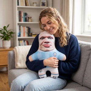 Pu&ograve; includere: Una bambola di peluche con un volto stampato realistico, che indossa una maglia da calcio azzurra e bianca. La bambola ha una testa rotonda e un'espressione sorridente. La bambola &egrave; tenuta da una persona.
