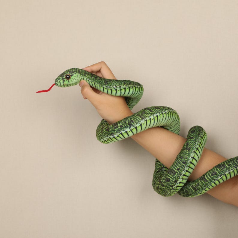 May include: A green and black plush snake toy with a red tongue. The snake is wrapped around an arm. The snake has a detailed pattern and is made of soft material. The background is a solid beige color.