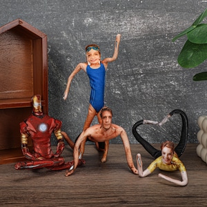 Puede incluir: Colección de muñecos de tela con varias figuras. Un muñeco está vestido como Iron Man, otro como nadador con un traje de baño azul, y otros en varias poses. Los muñecos están colocados sobre una superficie de madera.