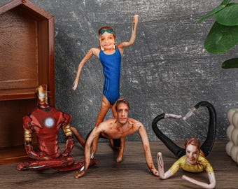 May include: A collection of fabric dolls featuring various figures. One doll is dressed as Iron Man, another as a swimmer in a blue swimsuit, and others in various poses. The dolls are arranged on a wooden surface.