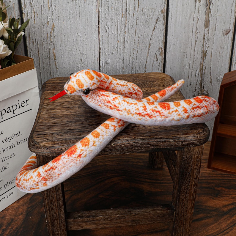 May include: A plush snake toy with a white body and orange and red markings. The snake has a red tongue and black eyes. It is resting on a small wooden stool.