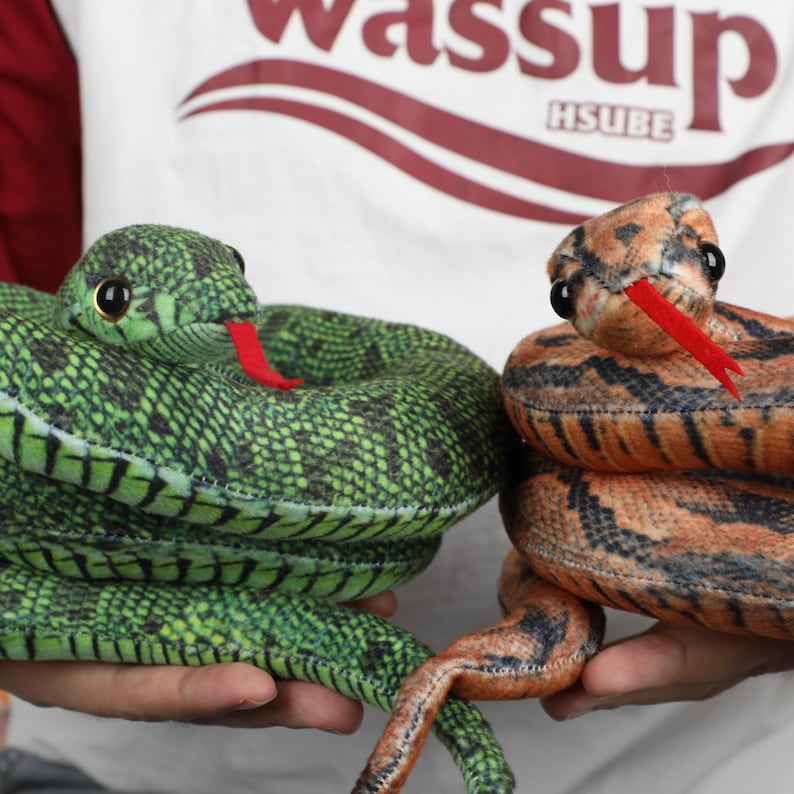 May include: Two plush snake toys. One is green with black markings and a red tongue. The other is brown and orange with black markings and a red tongue. Both have black eyes and are coiled.