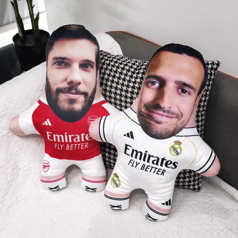 May include: Two plush dolls with faces printed on them, dressed in soccer jerseys. One doll wears a red and white Arsenal jersey with "Emirate Fly Better" printed on it. The other doll wears a white Real Madrid jersey with the same text.
