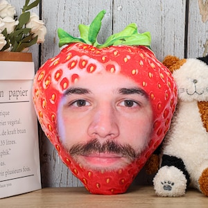 Peut inclure: Un coussin en forme de fraise rouge avec un visage de personne imprimé dessus. Le coussin a une tige et des feuilles vertes.