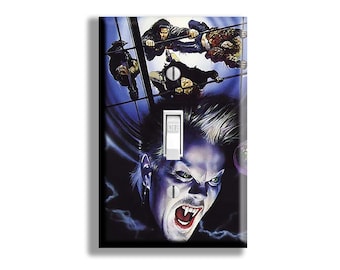 May include: A decorative light switch cover featuring a vampire with green eyes and an open mouth, with the word "OFF" on the switch. The background shows figures hanging from a structure, with a dark blue and purple color scheme.
