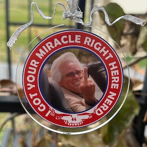 May include: A clear, round ornament with a silver ribbon for hanging. The ornament features a photo of a man in sunglasses giving the middle finger, surrounded by text that reads "I GOT YOUR MIRACLE RIGHT HERE NERD" in a red and white design.