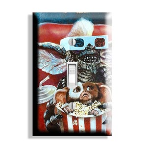 May include: A light switch cover featuring a colorful illustration of a Gremlin wearing 3D glasses and holding a popcorn bucket. The artwork includes a soda cup and a red and white striped popcorn container. The switch is in the "off" position.