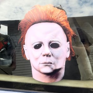 May include: A full-head mask featuring a pale, white-skinned human face with deep, dark, hollowed-out eye sockets and a neutral expression. The mask is topped with a thick, textured layer of bright orange hair that covers the crown and sides.