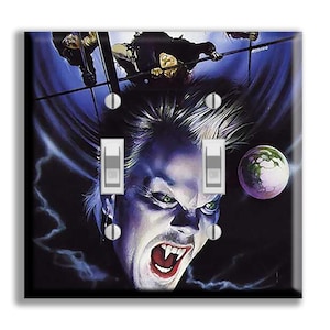May include: A decorative light switch cover featuring a vampire with fangs and green eyes, set against a dark blue and black background. The artwork includes figures and a glowing orb. The cover has two light switches.
