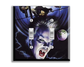 May include: A double light switch wall plate featuring a dark, dramatic illustration of a vampire with sharp fangs, green eyes, and spiky hair against a deep blue background with a glowing moon and figures hanging from a structure above.