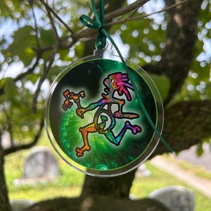 May include: A round, clear acrylic ornament featuring a colorful, stylized figure with a staff, set against a green and black background. The figure is in shades of orange, yellow, blue, and purple. A green ribbon is attached for hanging.