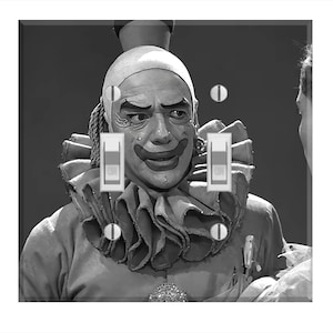 May include: A black and white light switch cover featuring a vintage image of a clown with makeup and a ruffled collar. The clown's face is the focal point, with the light switches integrated into the design.