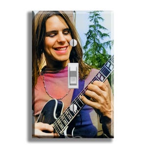 May include: A light switch cover featuring a smiling person playing a black electric guitar. The person is wearing a pink, purple, and blue shirt and a silver necklace. The background includes a tree and sky.