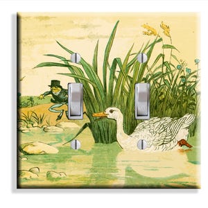 May include: A double light switch plate featuring a printed illustration of a white duck swimming in a pond near green reeds and a frog wearing a top hat and coat on the shore. The plate has two vertical toggle switches and four metal screws.