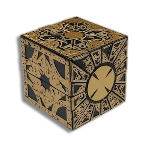 May include: A gold and black puzzle box with intricate geometric patterns. The cube-shaped box has a circular design on one side and a complex, multi-layered structure. The overall design is detailed and ornate.