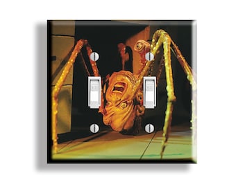 May include: A light switch cover featuring a grotesque creature with multiple legs and a screaming face. The artwork is in shades of orange, yellow, and brown, with two light switches and screws.