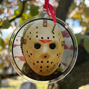 May include: A round, clear acrylic ornament featuring a close-up of a hockey mask with a red triangle on the forehead and a crack down the middle. The mask has black eye holes and numerous small holes. Red ribbon is attached for hanging.