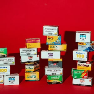 May include: A collection of various film boxes, including Kodak, Ilford, and Arista Edu brands, arranged against a red background. The boxes display text indicating film type, speed, and other specifications. The image showcases a variety of film options for photography enthusiasts.