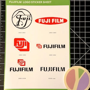 May include: A sticker sheet featuring various Fujifilm logos from different eras, including the years 1934-1960, 1960-1980, 1980-1992, 1985-1992, 1995-2006, and 2006-now. A round sticker with a colorful design is also present.