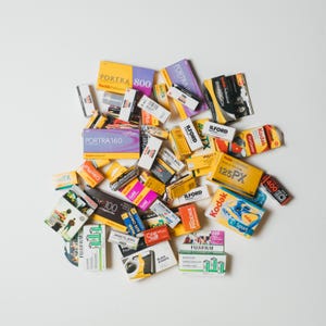 May include: A collection of various film rolls and boxes, including Kodak Portra 800, 160, and Gold, Ilford, and Fujifilm brands. The packaging features a range of colors, such as yellow, purple, and orange, and includes text indicating film type and speed.
