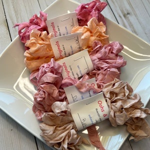 May include: A white square dish holds several bundles of ruffled silk ribbon in shades of pink, peach, and tan. Each bundle is labeled with a small tag that reads "Oohs & Originals". The ribbons are arranged in a visually appealing display.