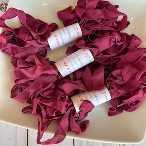 May include: Three spools of burgundy red fabric ribbon, each with a white label that reads "oohsahsooriginals" and "color: Plum" or "color: Blue".