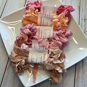 May include: A white ceramic dish holds several bundles of ruffled silk ribbon in shades of pink, peach, and tan. Each bundle is labeled with a small tag. The ribbon is arranged on a white surface.