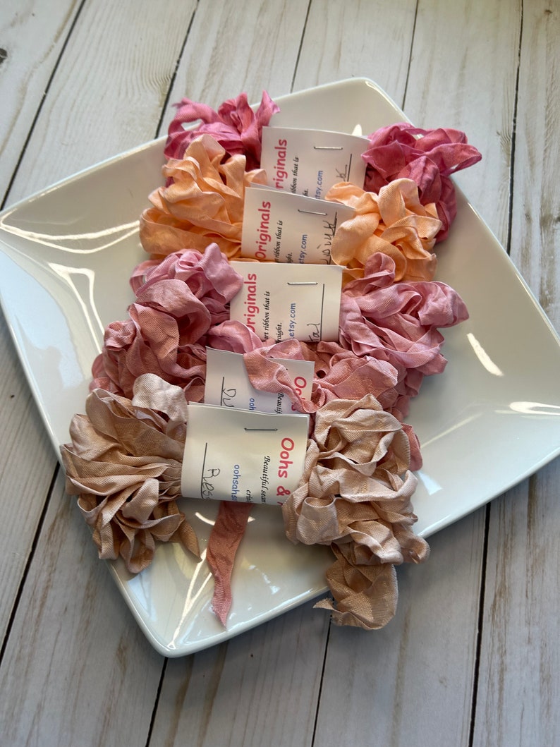 May include: A white ceramic dish holds several bundles of ruffled silk ribbon in shades of pink, peach, and tan. Each bundle is labeled with a small tag. The ribbon is arranged on a white surface.