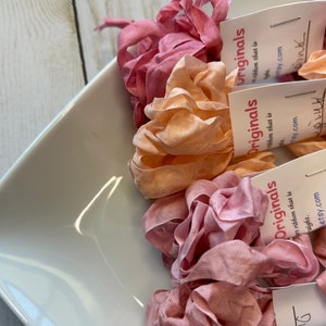 May include: A white square dish holds several bundles of pink and peach-colored ribbon. Each bundle is labeled with a small white tag that reads "Originals" and "art ribbon that is straight. etsy.com". The ribbons are arranged in a decorative manner.