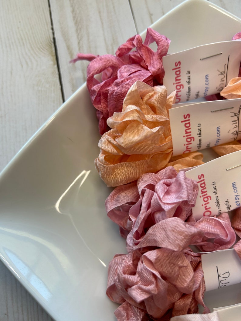 May include: A white square dish holds several bundles of pink and peach-colored ribbon. Each bundle is labeled with a small white tag that reads "Originals" and "art ribbon that is straight. etsy.com". The ribbons are arranged in a decorative manner.