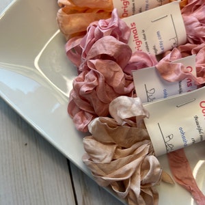 May include: A white rectangular dish filled with various shades of pink and peach-colored ribbon. The ribbon is arranged in ruffled, flower-like shapes. Small white tags with handwritten text are attached to some of the ribbon.