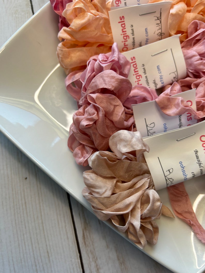 May include: A white rectangular dish filled with various shades of pink and peach-colored ribbon. The ribbon is arranged in ruffled, flower-like shapes. Small white tags with handwritten text are attached to some of the ribbon.