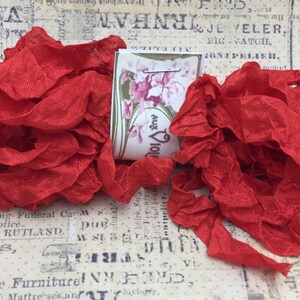 May include: Two spools of red ruffled fabric ribbon. The ribbon is a soft, crinkled texture. The spools are labeled "101 aux" and are sitting on a vintage printed paper background.