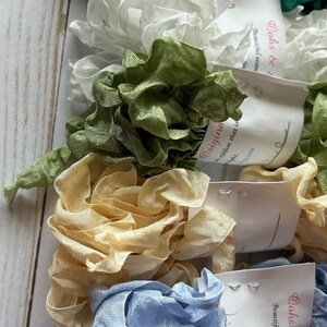 May include: A collection of ruffled ribbon in various colors, including green, white, light green, beige, and light blue. Each ribbon is bundled and tagged with a label that reads "Originals" and "Oohs & Beautiful seam". The ribbons are arranged on a light wood surface.