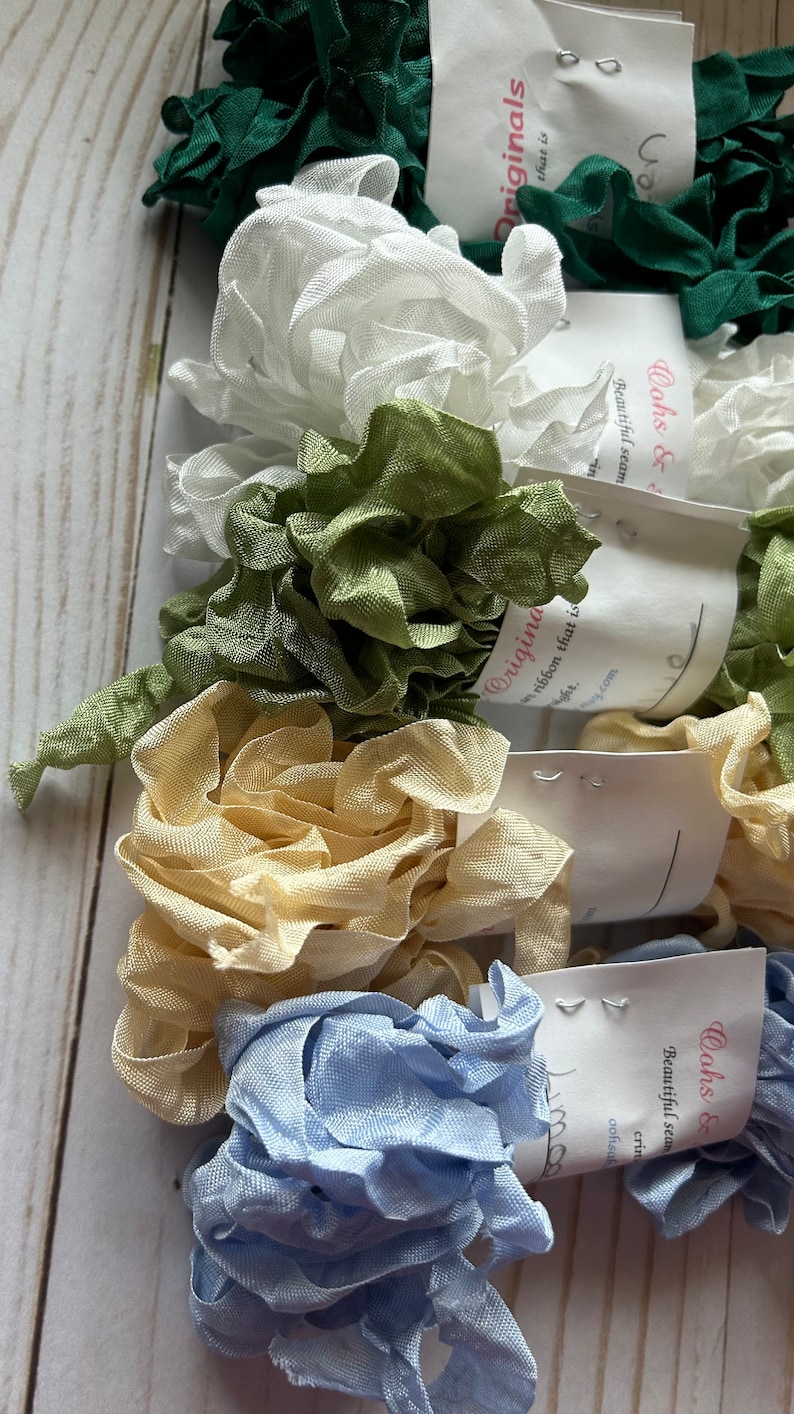 May include: A collection of ruffled ribbon in various colors, including green, white, light green, beige, and light blue. Each ribbon is bundled and tagged with a label that reads "Originals" and "Oohs & Beautiful seam". The ribbons are arranged on a light wood surface.