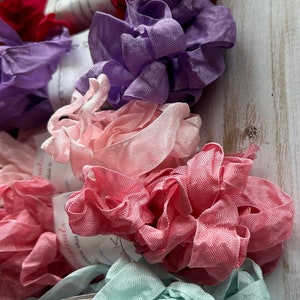 May include: A collection of colorful fabric ribbon rolls in shades of red, purple, pink, and teal. The ribbons are ruffled and textured, with small tags attached. The background is a light wood surface.