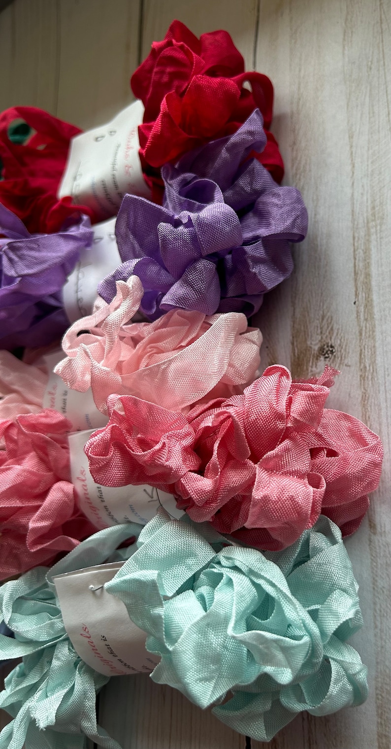 May include: A collection of colorful fabric ribbon rolls in shades of red, purple, pink, and teal. The ribbons are ruffled and textured, with small tags attached. The background is a light wood surface.