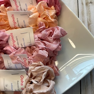 May include: A white, square-shaped dish filled with several spools of ruffled ribbon in shades of pink, peach, and beige. Each spool has a small tag with the text "Originals" and a website address.