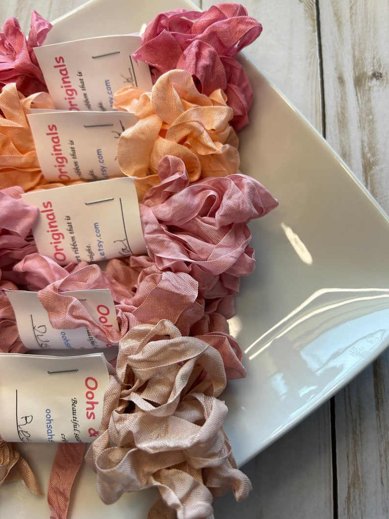 May include: A white, square-shaped dish filled with several spools of ruffled ribbon in shades of pink, peach, and beige. Each spool has a small tag with the text "Originals" and a website address.
