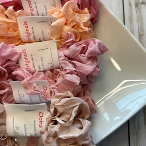 May include: A collection of ruffled silk ribbons in shades of pink, peach, and beige, displayed in a white, rectangular dish. Each ribbon bundle has a tag with the text "Oohs & Originals" and a website address. The ribbons are arranged in a visually appealing display.