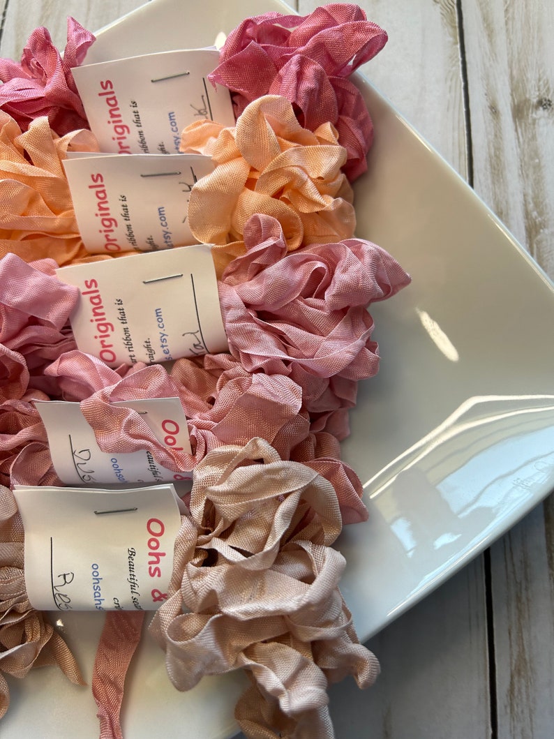 May include: A collection of ruffled silk ribbons in shades of pink, peach, and beige, displayed in a white, rectangular dish. Each ribbon bundle has a tag with the text "Oohs & Originals" and a website address. The ribbons are arranged in a visually appealing display.