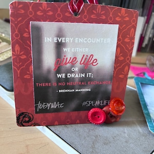 May include: Red and white card with a quote about giving life or draining it. The quote is by Brennan Manning. The card has a red ribbon and two red buttons attached to it. The card says "#SPEAKLIFE" and "TOBYMAC".