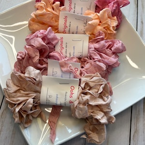 May include: A collection of ruffled silk ribbons in shades of pink, peach, and beige, displayed on a white square plate. Each ribbon is bundled and tagged with a small white label. The ribbons are arranged on a light wood surface.